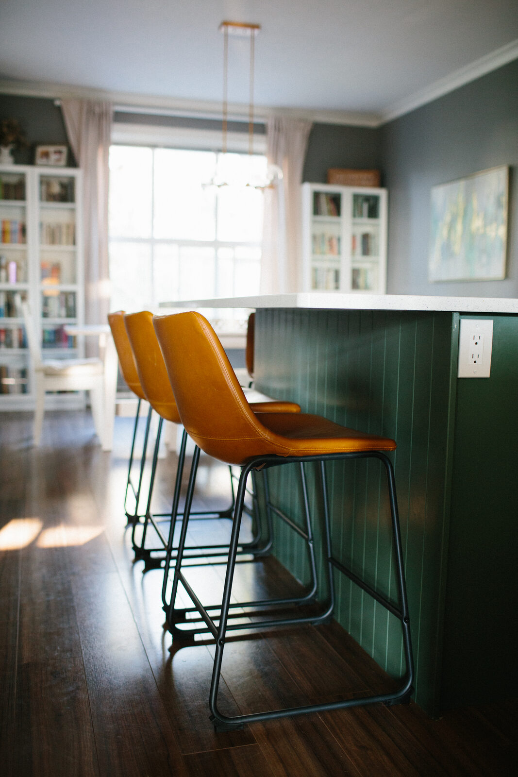 Green V-groove panelled kitchen island with a white slab top and leather counter stools on dark plank flooring